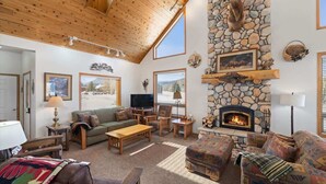 A cozy living room with a stone fireplace, wooden ceiling, and large windows.