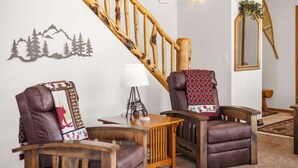 A cozy living room with a wooden staircase, two leather armchairs, a small table with a lamp, and a wall-mounted mirror.