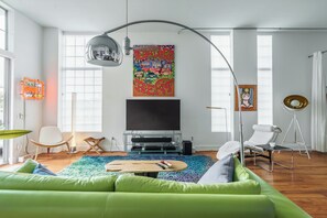 A modern living room with a green sofa, a flat-screen TV, a colorful rug, and a large window.