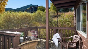 Une terrasse en bois, agrémentée de chaises et d’une table en osier, qui offre une vue sur une zone boisée avec une montagne en arrière-plan.