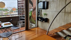 Un espace de vie moderne doté d’un plancher en bois, d’un coin repas avec une table et des chaises, d’un téléviseur fixé au mur et d’un escalier en colimaçon menant à un espace extérieur offrant une vue sur la ville.