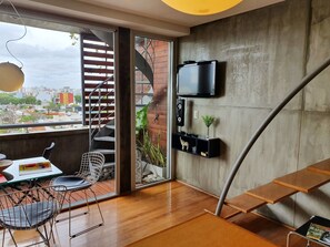 A modern living space with a wooden floor, a dining area with a table and chairs, a TV mounted on the wall, and a spiral staircase leading to an outdoor area with a city view.