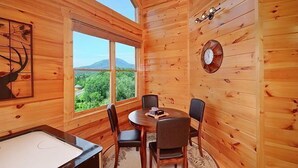 Interior d'una cabana de fusta amb taula rodona, quatre cadires, un rellotge i una finestra amb vistes a vegetació i muntanyes.