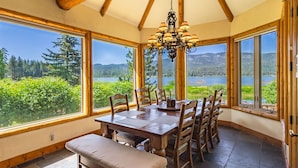 A dining area with a wooden table and chairs, large windows with a view of a lake and forest, and a chandelier.