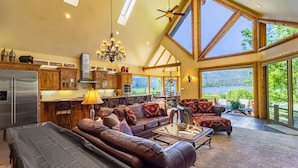 A spacious living area with a large brown leather sofa, a coffee table, and a dining table with chairs. There is a kitchen with wooden cabinets and a stainless steel refrigerator. The room has large windows offering a view of the outdoors.