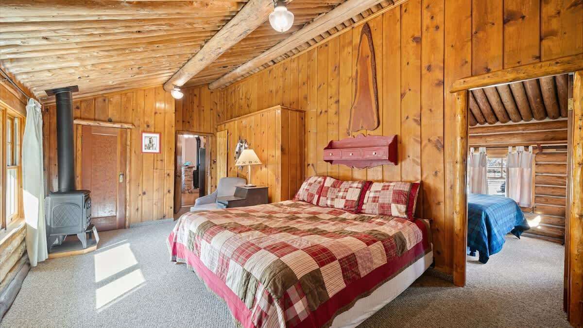 A cozy wooden cabin bedroom with a bed, a dresser, a lamp, and a wood-burning stove.