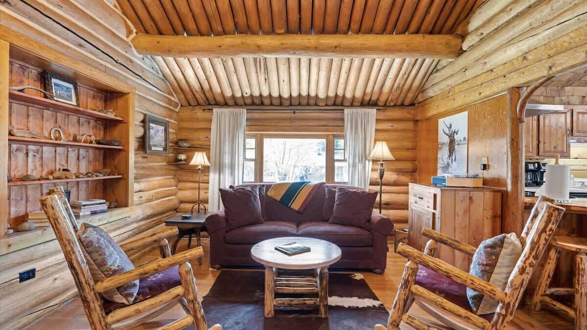 L’intérieur d’une cabane en rondins, avec un plafond en bois, un canapé, deux fauteuils, une table basse et un coin cuisine.
