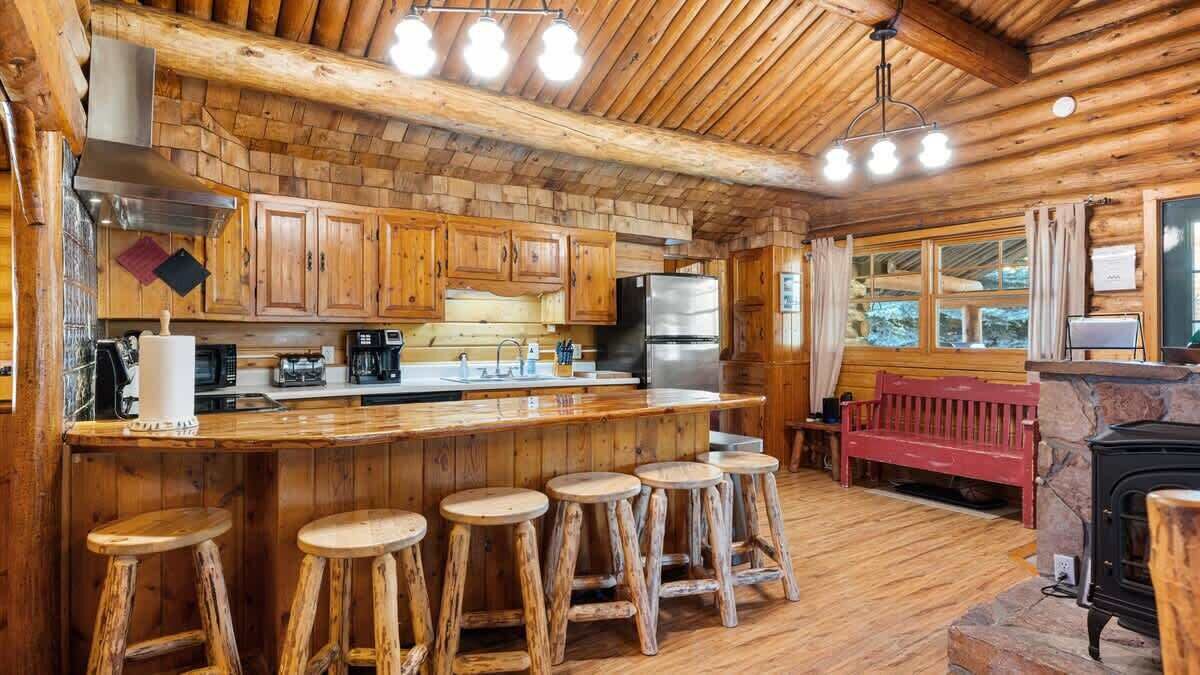 A rustic cabin-style kitchen with wooden cabinetry, a central island with stools, a red bench, and a wood-burning stove.