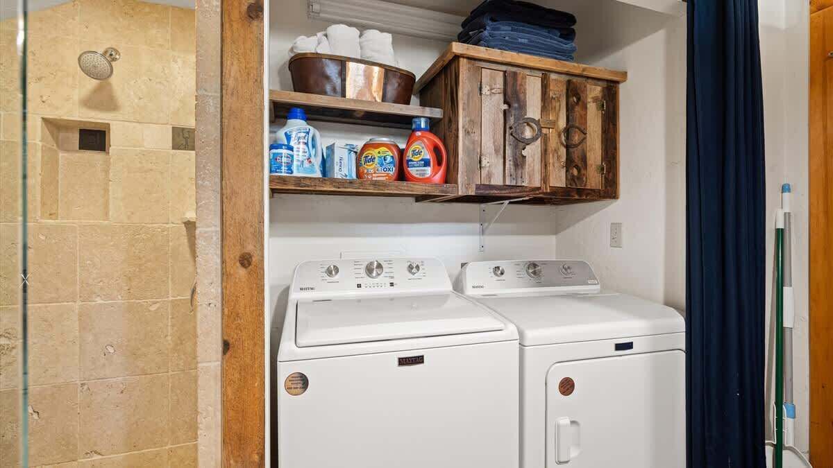 Une buanderie équipée de deux machines à laver et de deux sèche-linge à chargement frontal, tous deux de couleur blanche, une étagère en bois au-dessus pour ranger les produits de lessive, et un espace douche avec un mur en pierre.