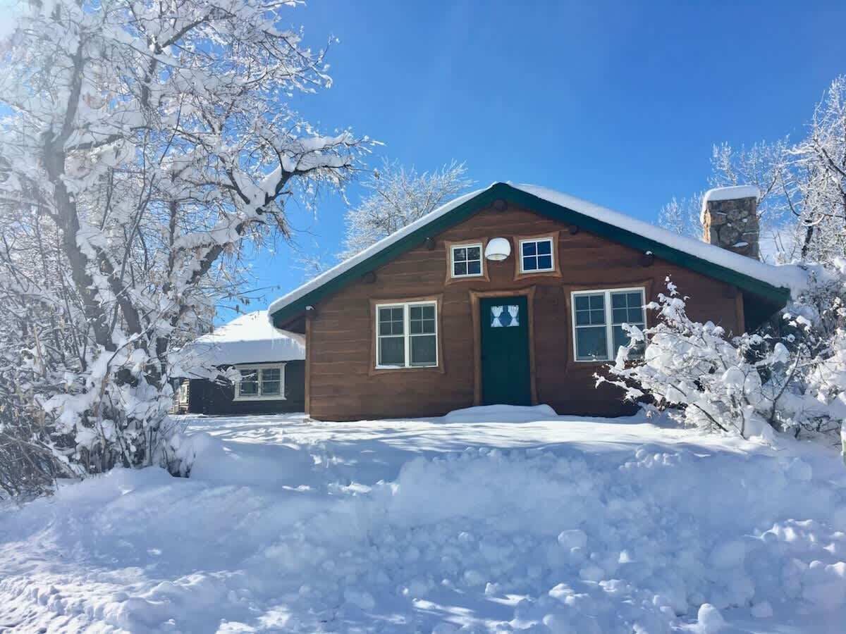 A wooden cabin with a green door, surrounded by snow-covered trees and a clear blue sky.