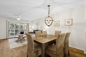 A dining area with a wooden table and wicker chairs, a chandelier, and a view of the outdoors through large windows.