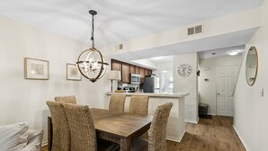 A dining area with a wooden table and wicker chairs, a kitchen in the background, and a clock on the wall. - 307 Sailmaker in Summer Beach (Fernandina Beach)
