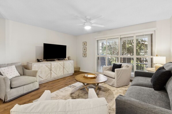 A living room with a flat-screen TV, white furniture, a ceiling fan, and large windows with blinds.