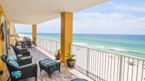 Un balcón con muebles de mimbre y plantas en macetas, con vista a una playa con gente y un cielo despejado.