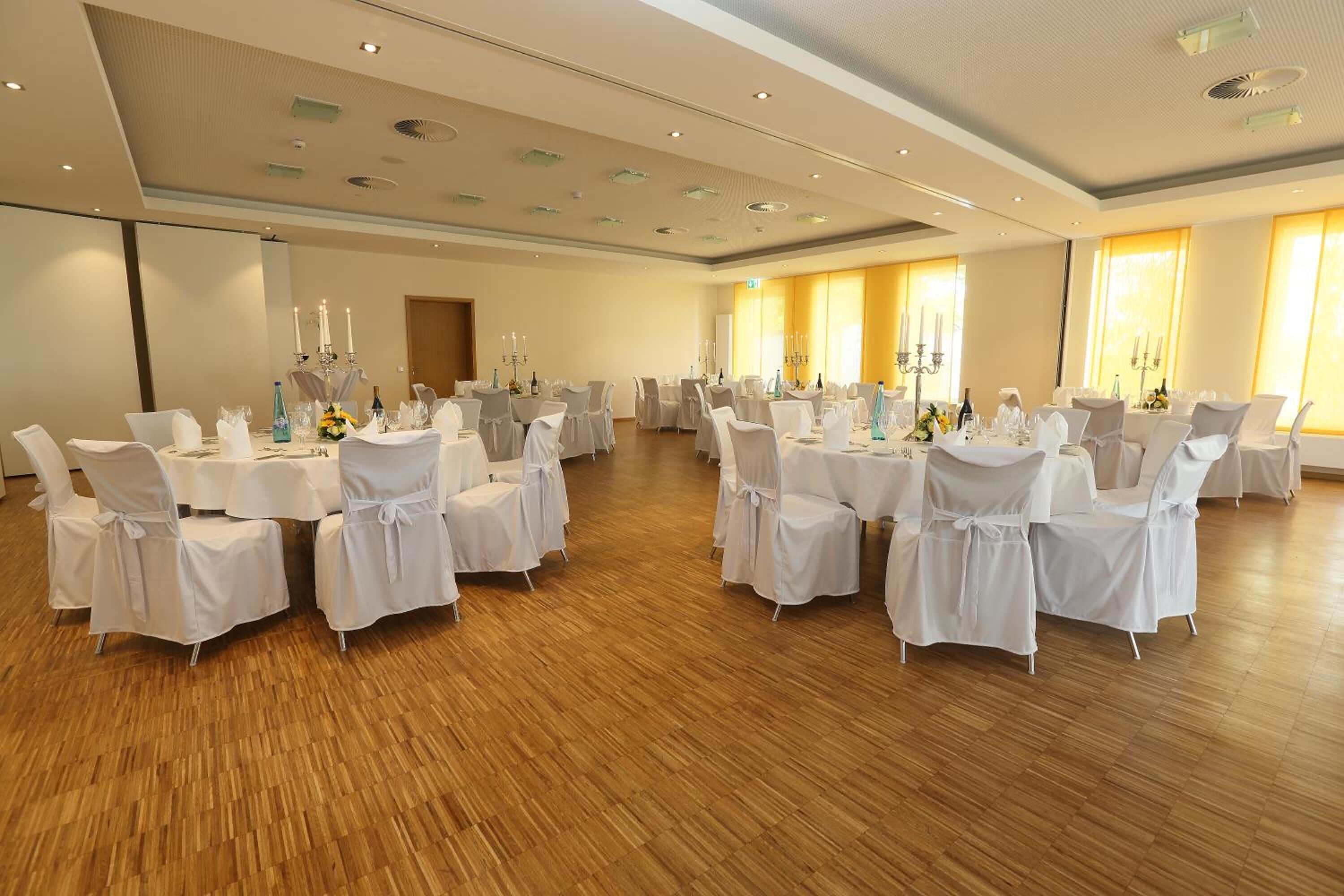 A large conference room with round tables and white chairs, wooden flooring, and large windows with curtains.