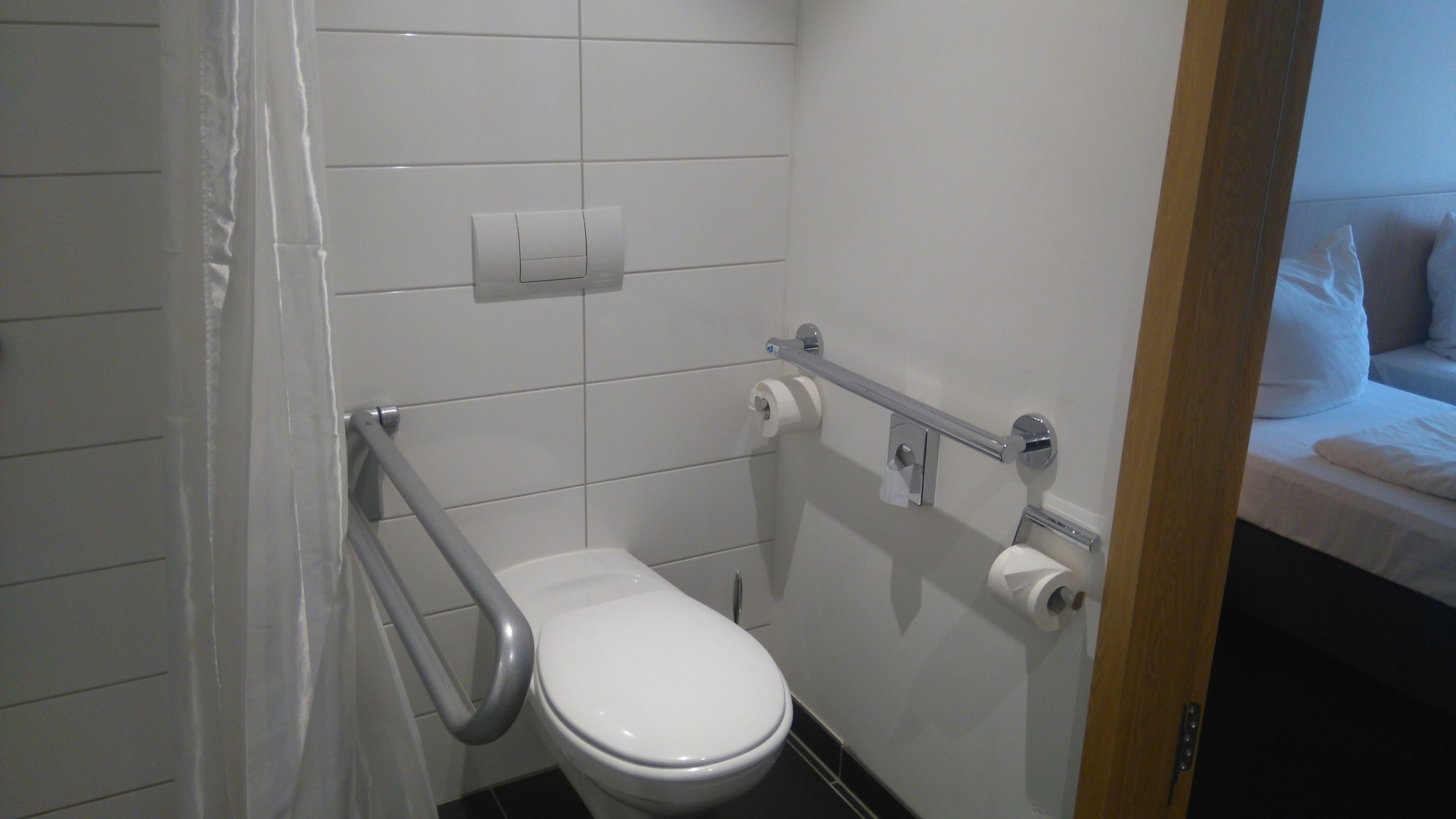 A small, white bathroom with a toilet, a handrail, and a towel dispenser.
