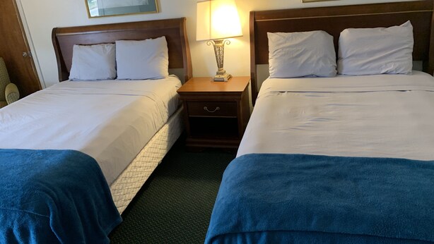 Habitación de hotel con dos camas, cada una con sábanas blancas y cabeceros de madera. Entre las camas hay una mesita de noche con una lámpara y un cajón pequeño.