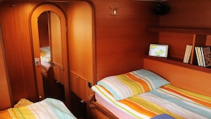 A compact room with two beds, wooden walls, a small shelf with books, and a tablet on the wall. - iCan Sailing Experience (Turneffe Islands)