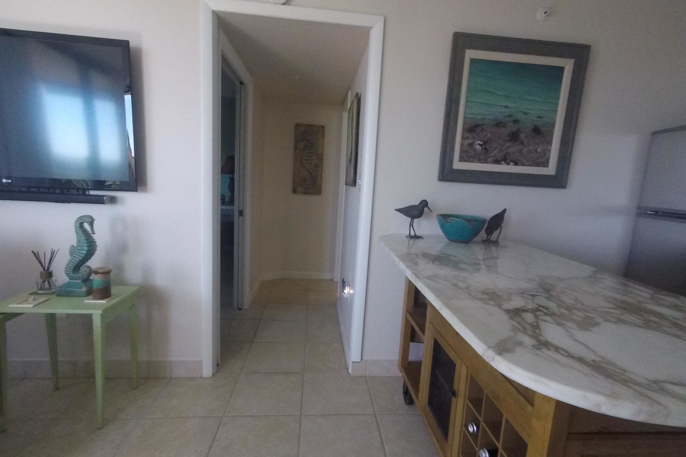 A kitchen with a marble countertop, a flat-screen TV, a small table with decorative items, a framed painting, and a bird figurine.