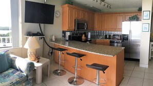 A modern kitchen with wooden cabinets, a stainless steel refrigerator, a microwave, and a built-in oven. There are three bar stools and a small island with a sink. A flat-screen TV is mounted on the wall above the kitchen counter. - Lovers Key Beach Club by Lovers Key Vacations (Fort Myers Beach)