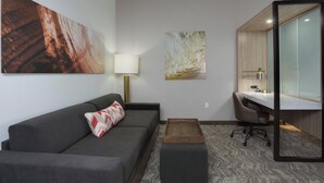 A modern hotel room with a gray sofa, a desk with a chair, a large mirror, and two abstract wall art pieces. - SpringHill Suites by Marriott Fort Lauderdale Miramar (Miramar)