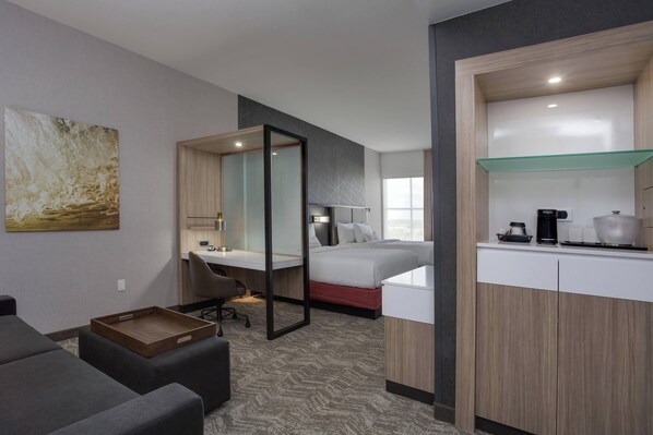 Habitación de hotel con cama, escritorio, silla, mesita pequeña, estante de vidrio y un cuadro en la pared.