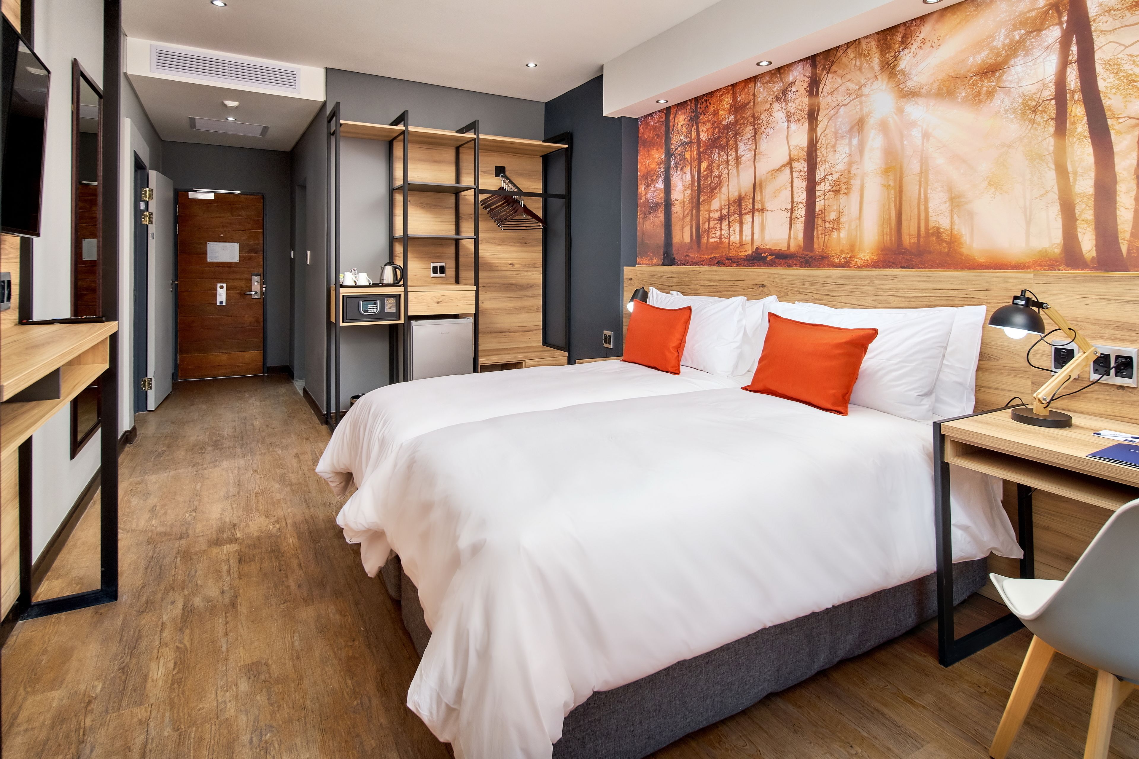 A modern hotel room with a large bed, a desk, a chair, a wardrobe, and a painting of a forest.
