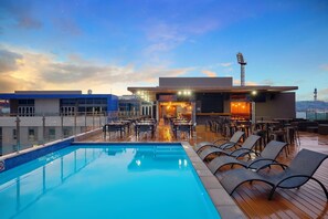 Un área de piscina en la azotea con sillones de descanso, un bar y vistas del horizonte de la ciudad.