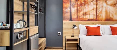 A modern hotel room with a bed, a nightstand, a desk, a mini-fridge, and a wall-mounted shelf.
