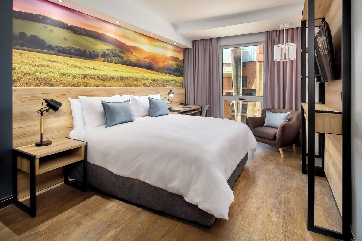 Ein modernes Hotelzimmer mit einem großen Bett, einem Schreibtisch, einem Sessel und einem an der Wand befestigten Fernseher.