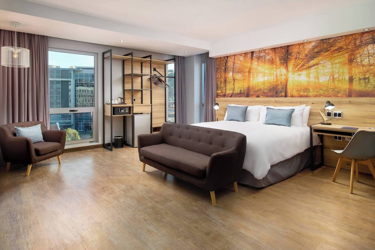 Ein modernes Hotelzimmer mit einem großen Bett, einer Couch, einem Schreibtisch und Blick auf die Stadt durch ein großes Fenster.