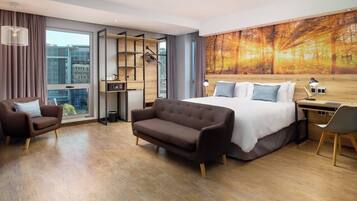 A modern hotel room with a large bed, a sofa, a desk, and a view of the city through a large window.