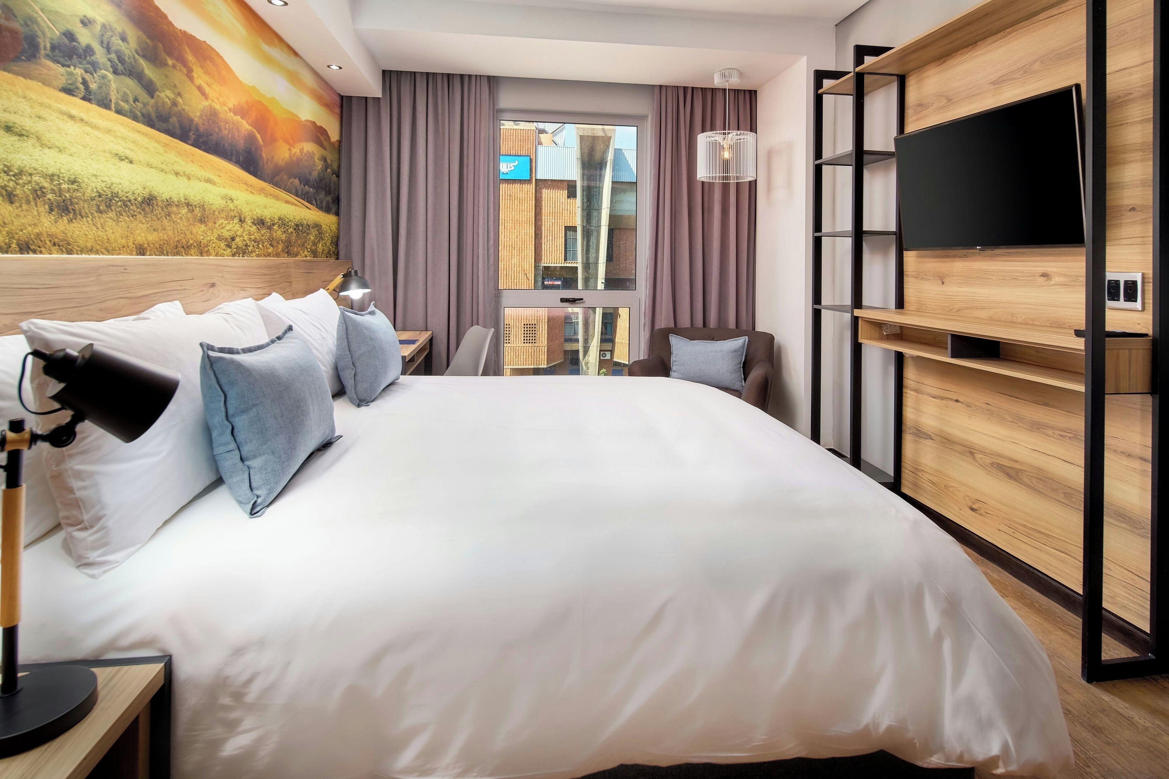 Ein Hotelzimmer mit einem großen Bett, einem Fernseher, einem Schreibtisch und Blick auf die Stadt durch das Fenster.
