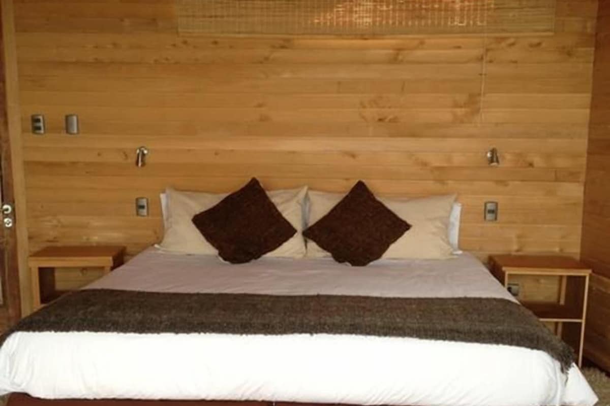 Quarto com painéis de madeira, uma cama, duas mesas de cabeceira e uma janela com persiana mosquiteira.