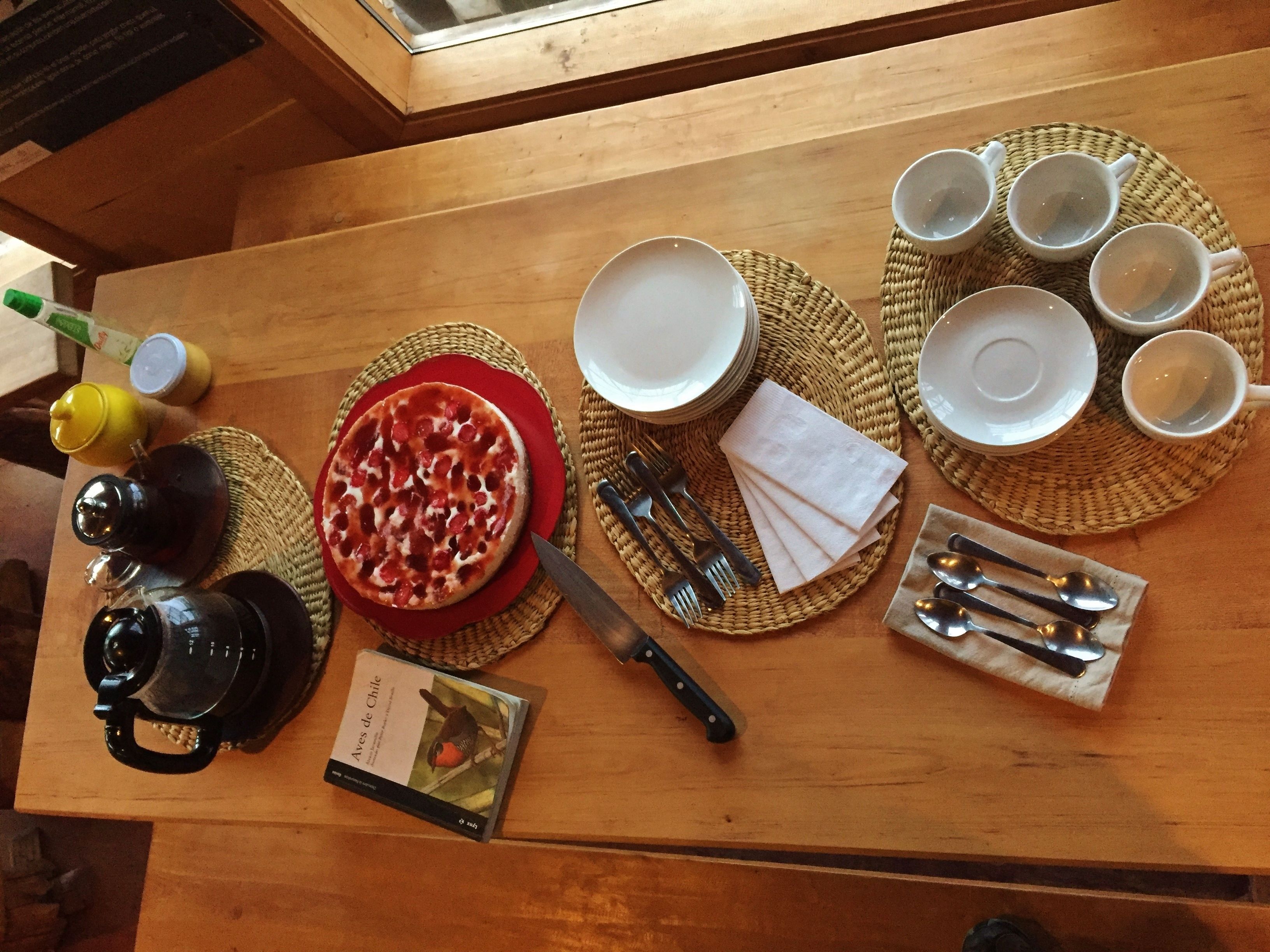 Ett träbord med en pizza, tallrikar, koppar, bestick, en bok och en kaffebryggare.