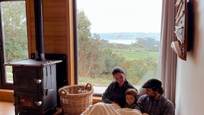 A cozy room with a wood-burning stove, a large window with a view of greenery, and a comfortable seating area. - Refugio Pullao (Castro)