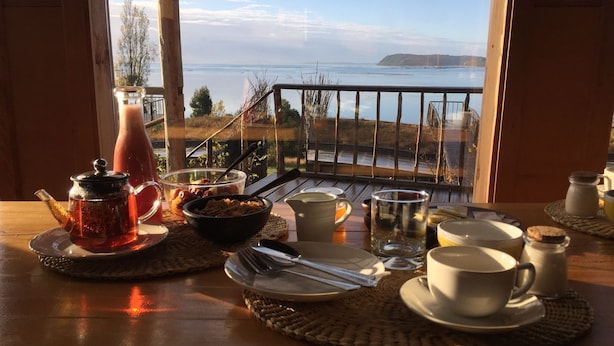 A breakfast table with tea, cereal, and cups, overlooking a scenic view of the sea and hills. - Refugio Pullao (Castro)