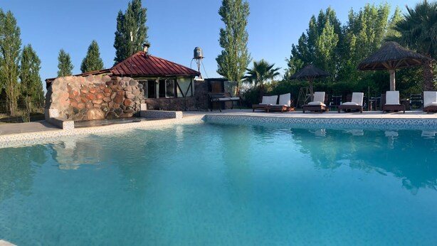 A clear blue pool with lounge chairs and umbrellas, surrounded by trees and a stone building with a red roof. - Cabañas Solitudine (Las Compuertas)