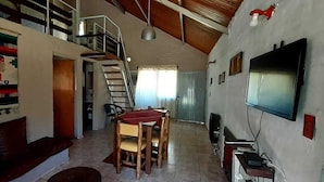 A spacious living area with a high ceiling, a staircase, a dining table, and a television mounted on the wall. - Cabañas Solitudine (Las Compuertas)