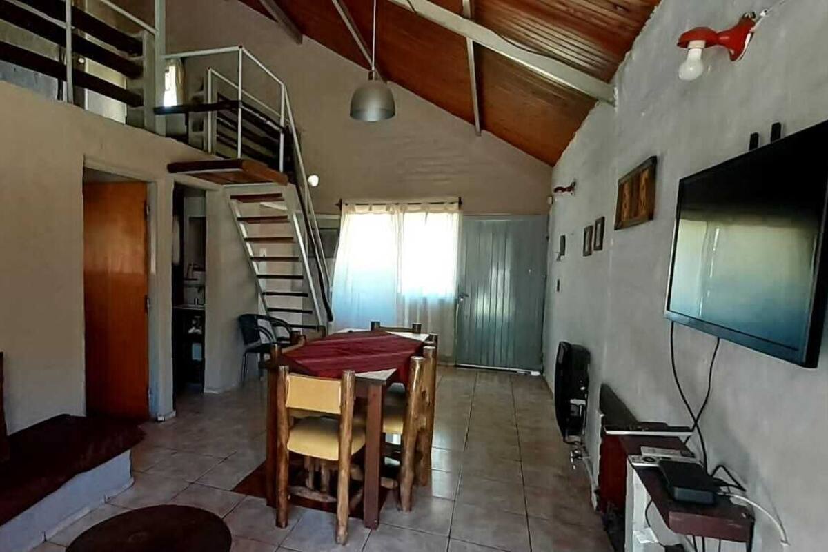 A spacious living area with a high ceiling, a staircase, a dining table, and a television mounted on the wall.