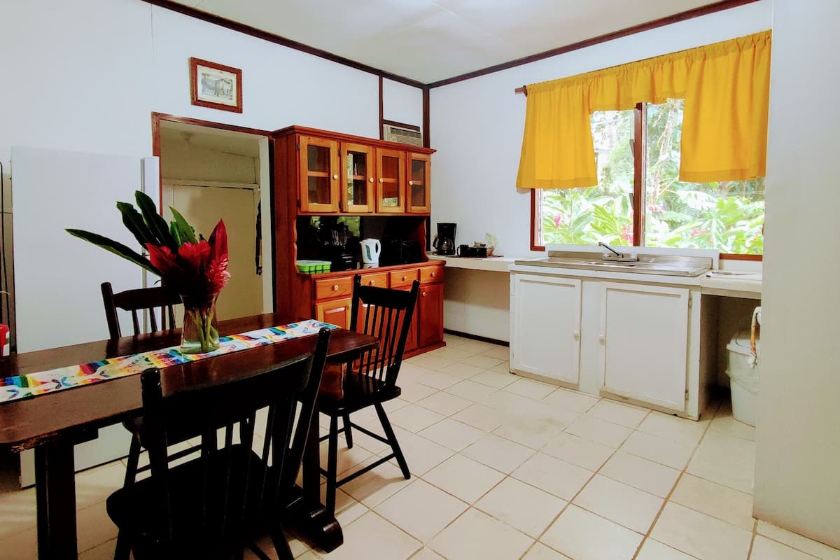 A compact kitchen with wooden cabinets, a dining table with chairs, and a window with yellow curtains.