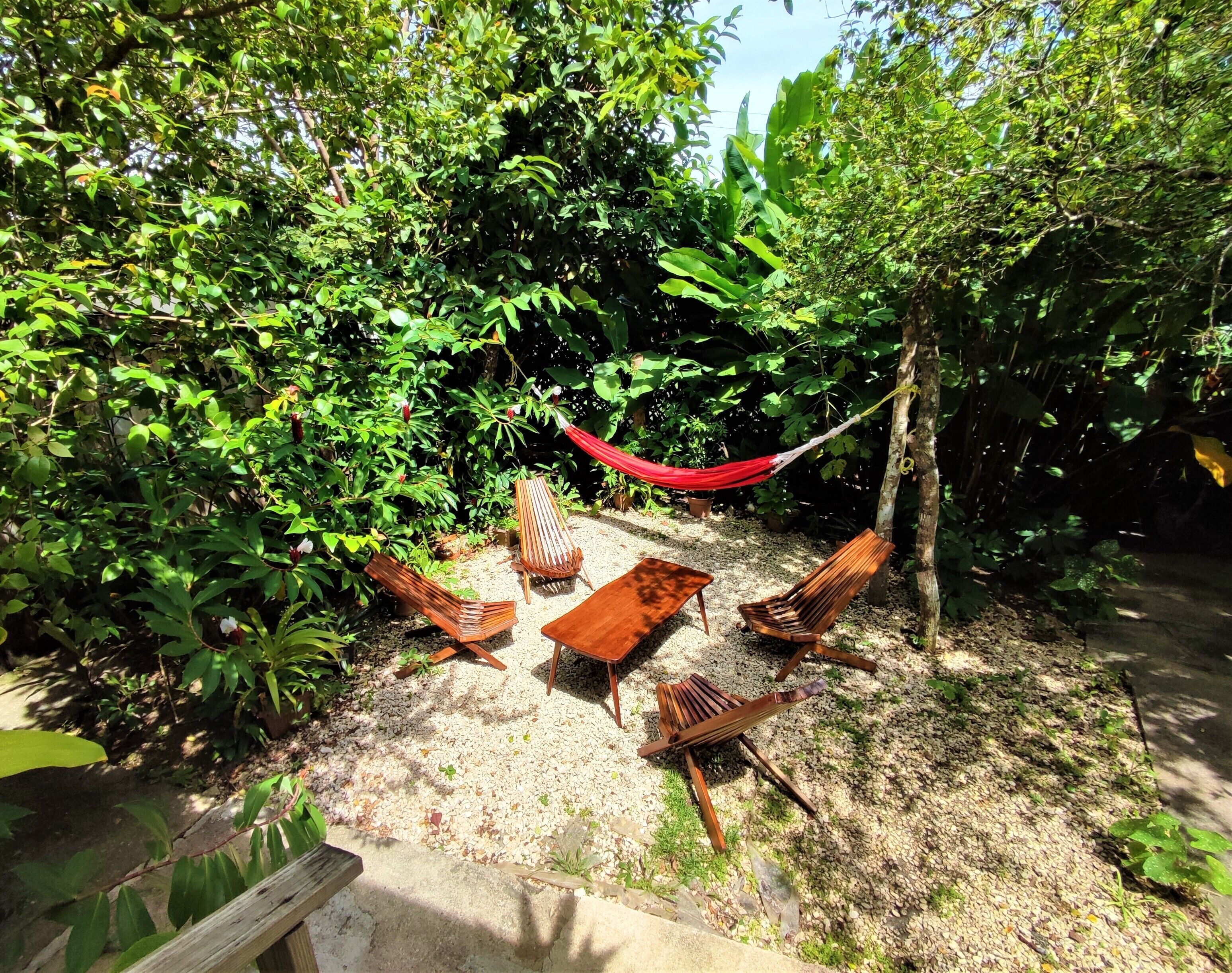 A garden area with a wooden table and four chairs, surrounded by dense foliage and a red hammock.