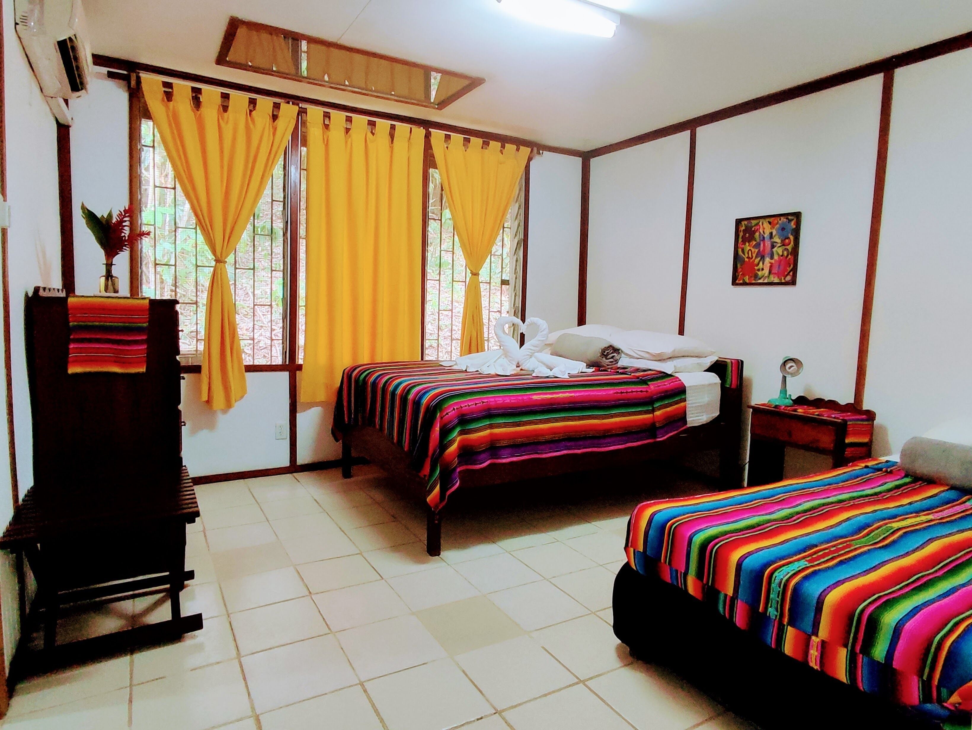 A room with two beds, a wooden bench, a small table with a lamp, and a window with yellow curtains.