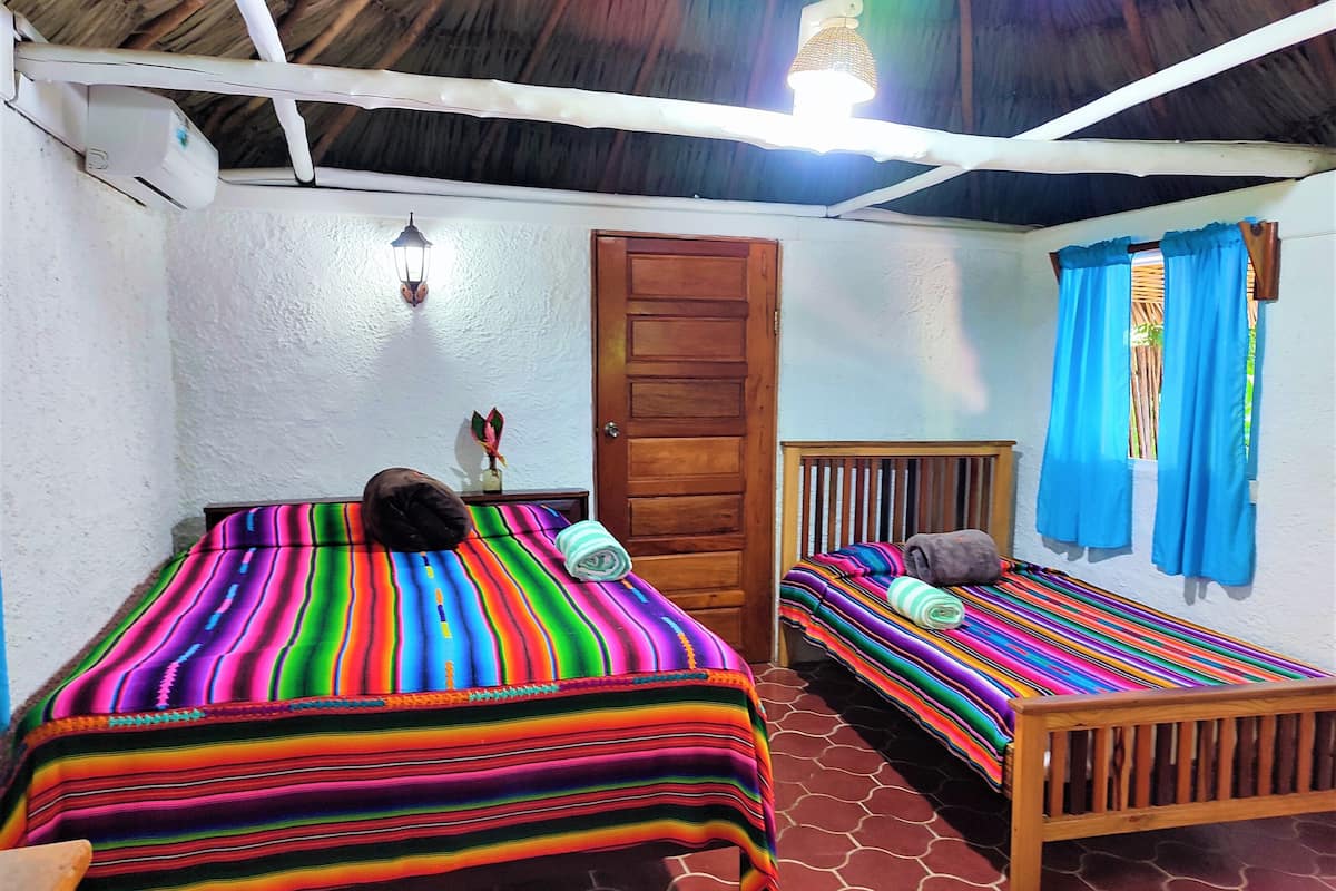 A room with two beds, a wooden door, a wall-mounted light fixture, and a window with blue curtains.