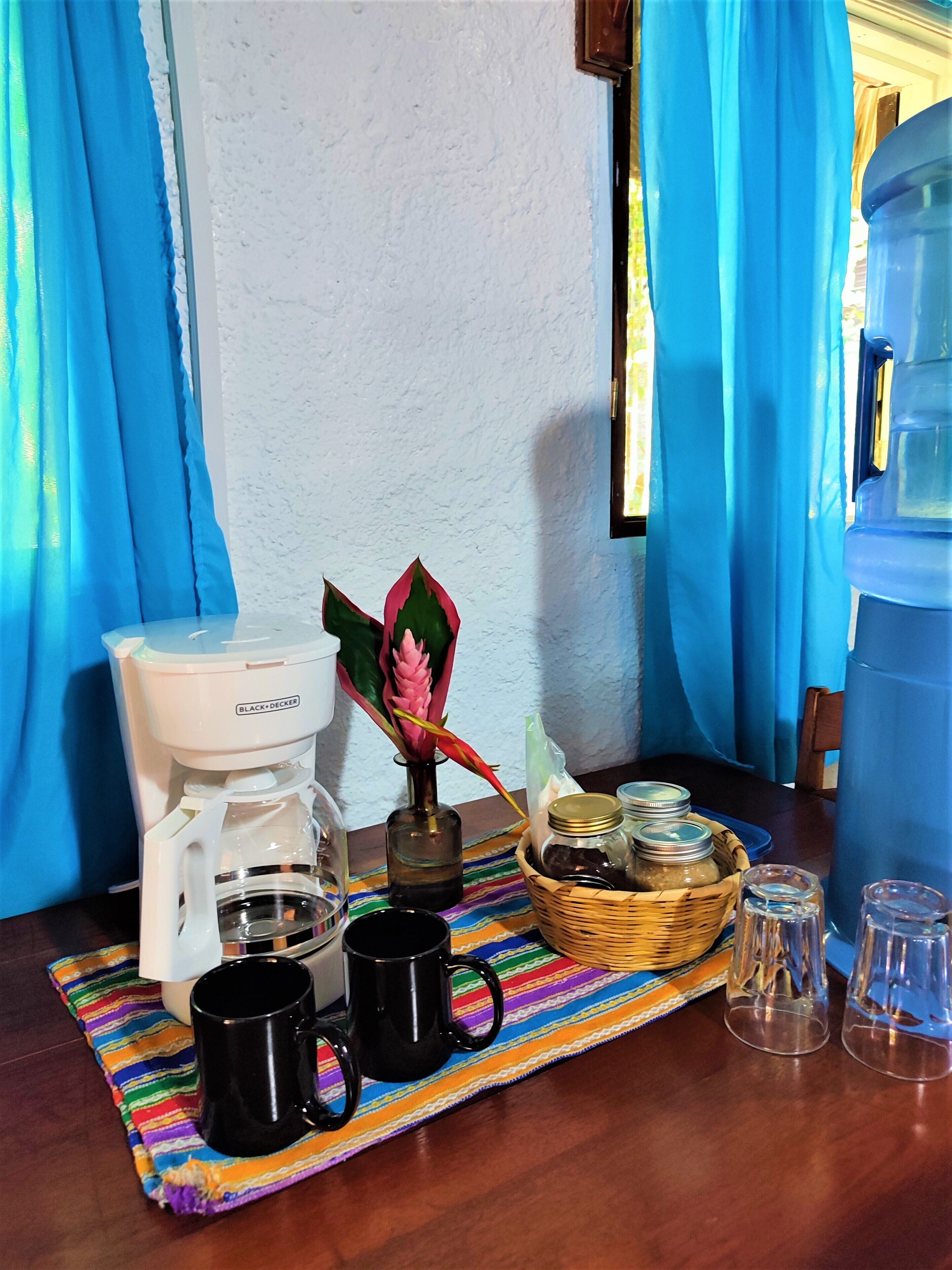 Uma estação de café com uma cafeteira, duas canecas pretas, uma cesta de vime com potes e um dispensador de água.