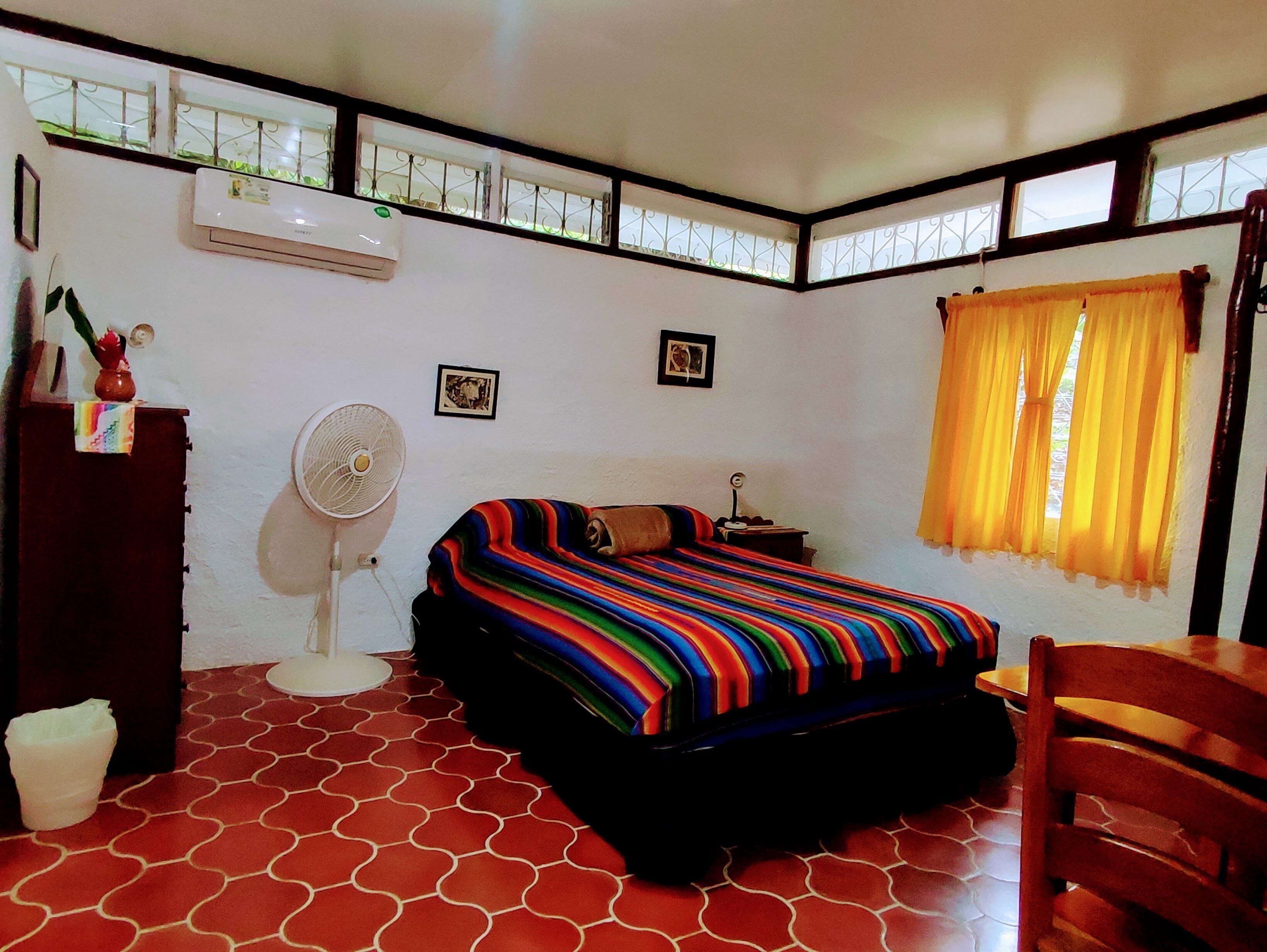 Um quarto com uma cama, um ventilador, uma janela com cortinas e um radiador.
