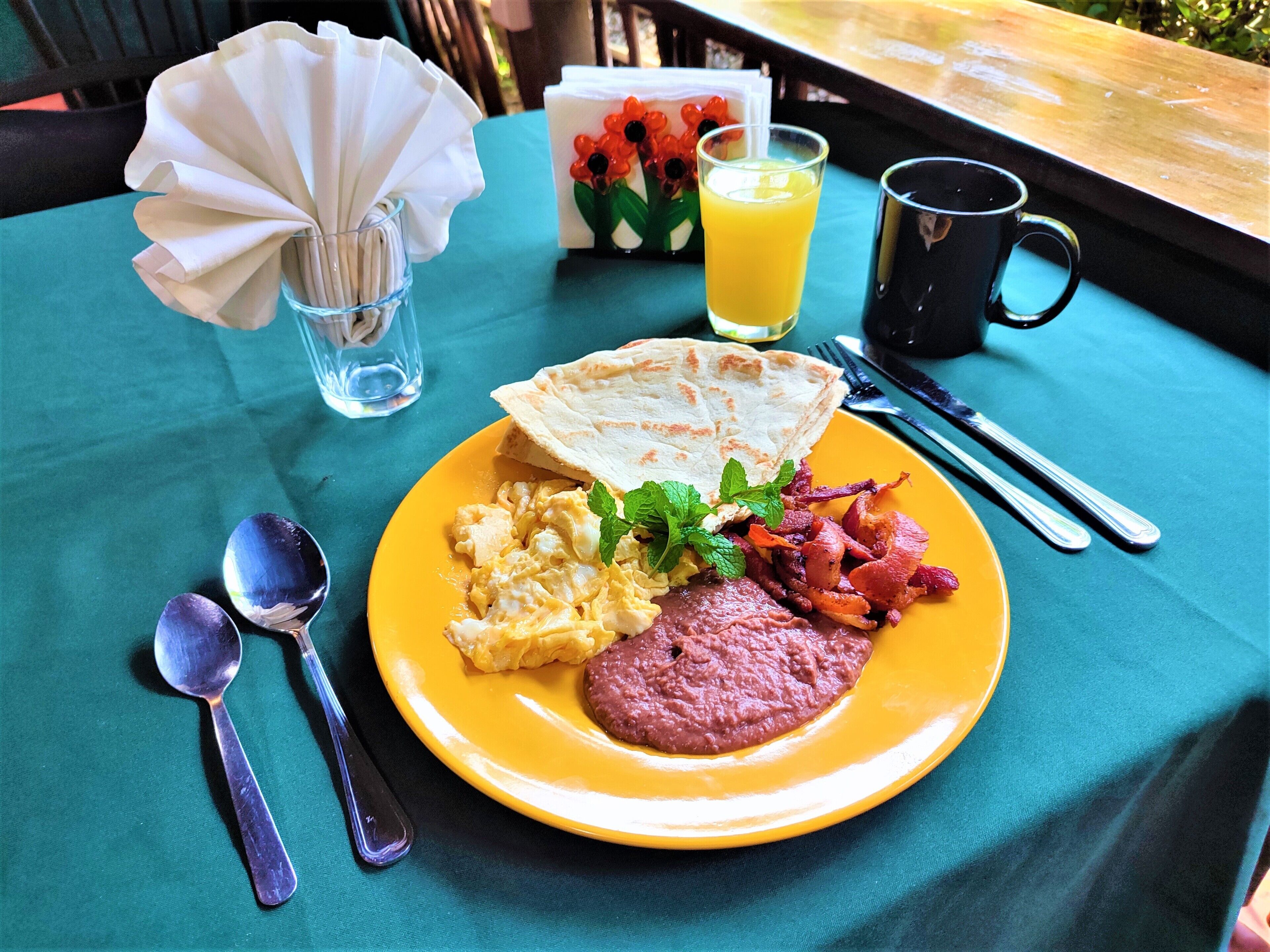 A breakfast plate with scrambled eggs, bacon, and toast on a yellow plate, accompanied by a glass of orange juice, a black mug, and cutlery on a teal tablecloth.