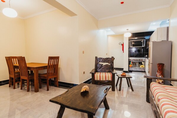 A living room with a dining table, chairs, a sofa, a small table with a decorative item, and a kitchen area with appliances. - Mountain View Hotel & Apartment (Kigali)