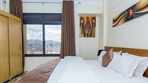 A hotel room with a large bed, a wooden wardrobe, a city view through the window, and a piece of abstract art on the wall.