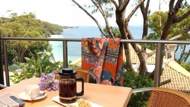 Ein Tisch mit Kaffeekanne, Tassen und einer Speiseplatte, vor einem Balkon mit Blick auf das Meer und Bäume.